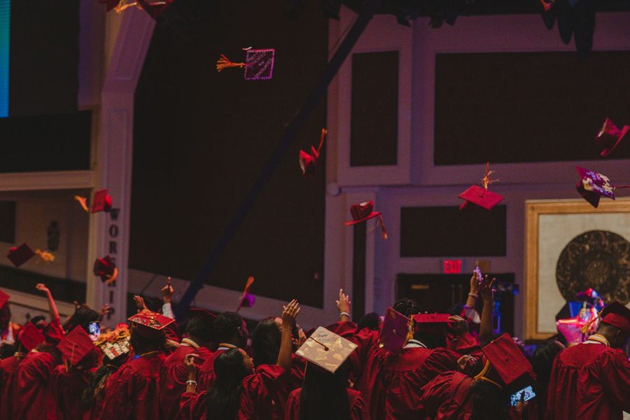 Graduates in red gowns throwing caps at an indoor graduation ceremony